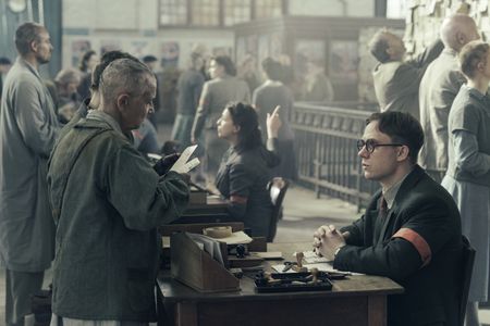 A SMALL LIGHT - Refugees examine photos of Anne and Margot Frank as Jan Gies, played by Joe Cole, looks on as seen in A SMALL LIGHT. (Credit: National Geographic for Disney/Dusan Martincek)