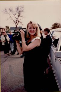 Jodi Huisentruit standing next to a limo in black dress, smiling and holding a camera on her shoulder.